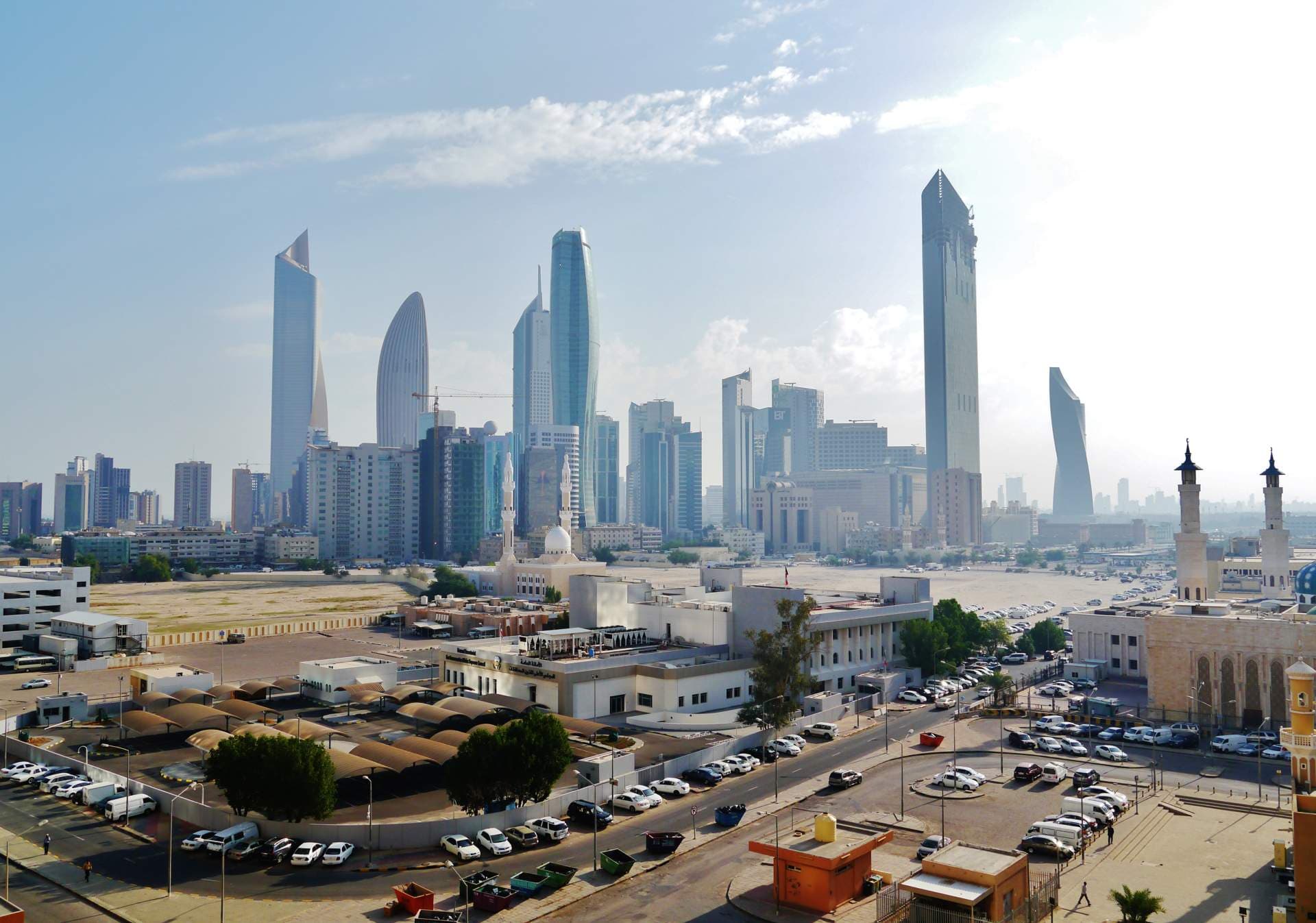 Aerial view of Kuwait City skyline with modern skyscrapers, mosques, and urban infrastructure under a partly cloudy sky.