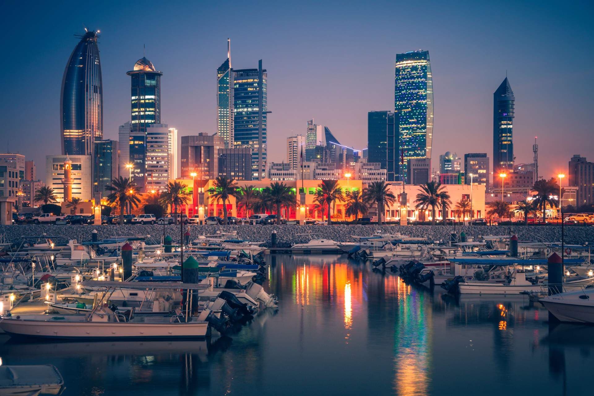 Kuwait City skyline at dusk with waterfront marina, boats, palm trees, and illuminated high-rises reflecting on the water.
