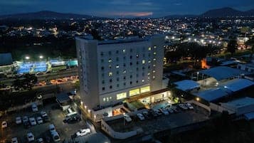 Aerial night view of a multi-story hotel building with a lit parking lot, overlooking a sprawling cityscape and distant mountains.
