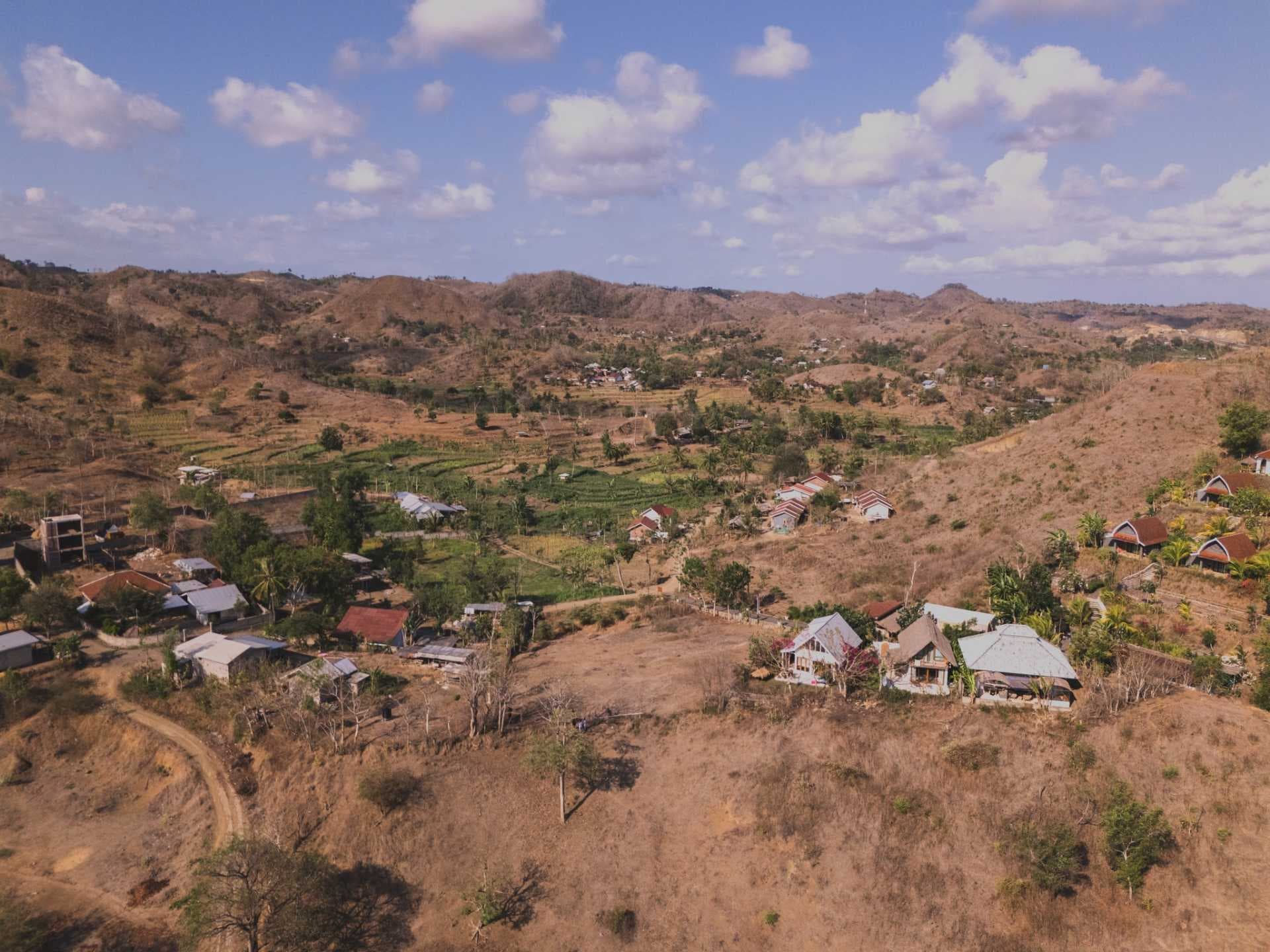 Aerial view of a rural village with simple homes and terraced fields on dry hills under a partly cloudy sky.