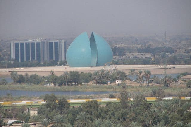 Wide hazy landscape view of Al ‘Amārah featuring a large blue dome-shaped structure, a modern building, palm trees, and the Tigris River.