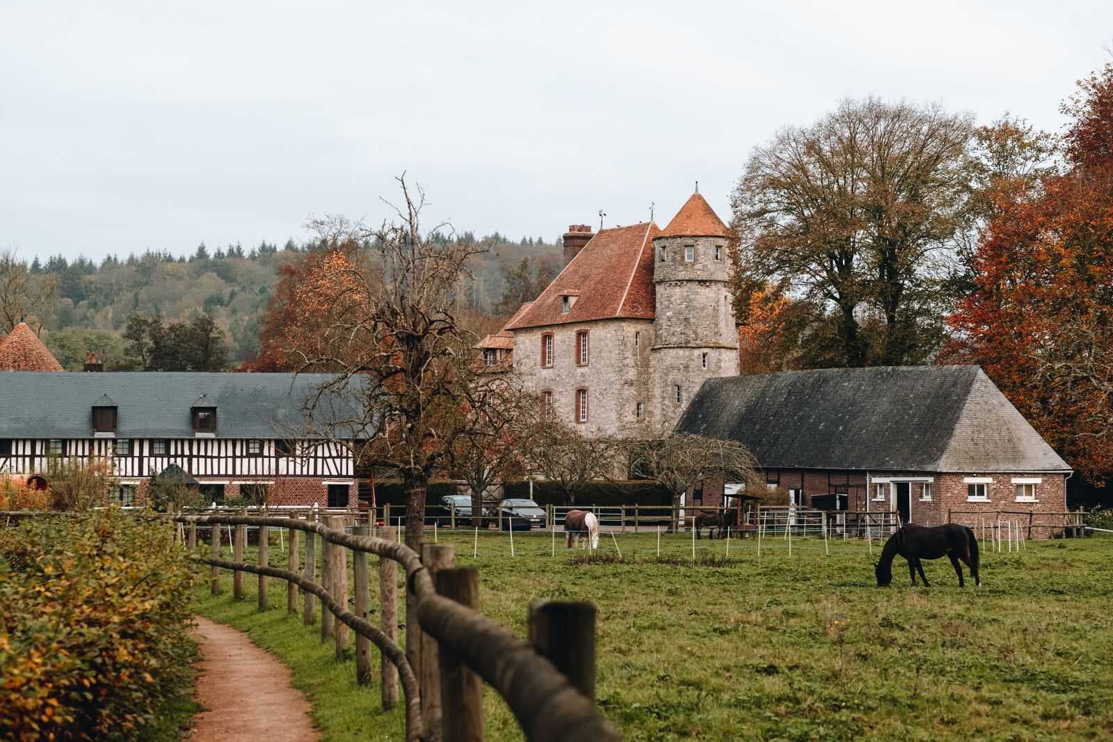 Rural estate with stone tower, timber-framed buildings, fenced pasture, grazing horses, and a dirt path under overcast skies near Roncherolles-sur-le-Vivier, Fr