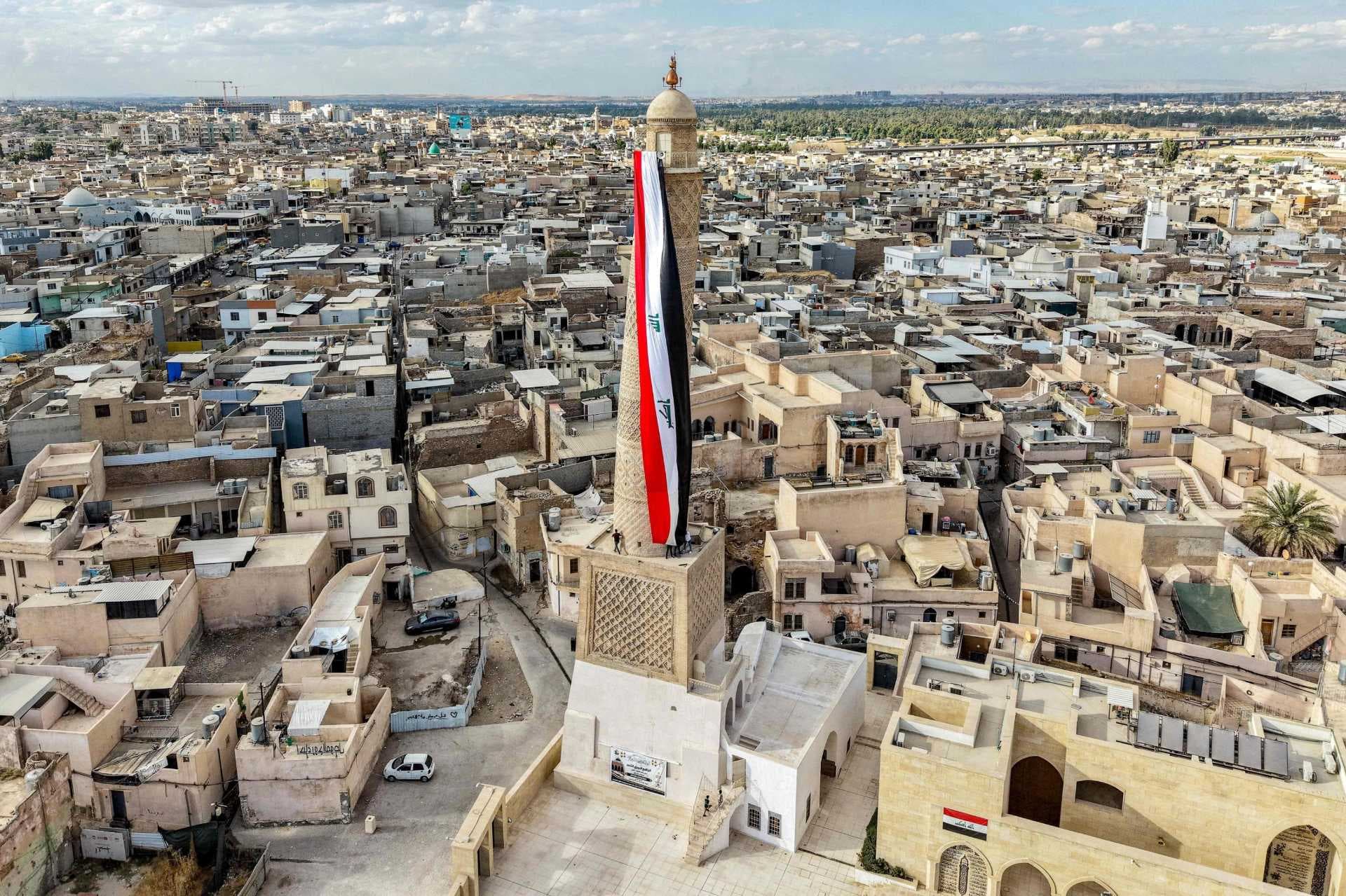 Aerial view of Mosul cityscape with a mosque minaret flying the Iraqi flag, surrounded by dense urban buildings.