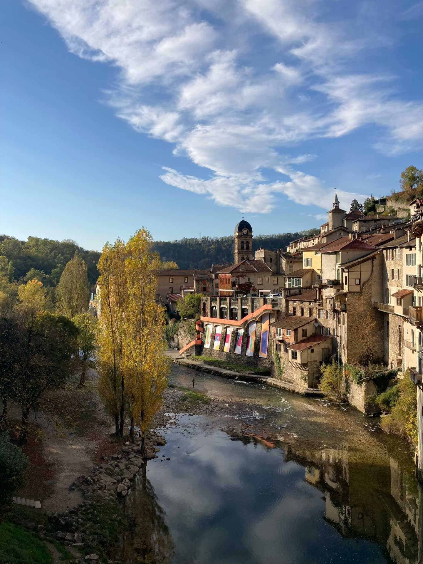 Elevated exterior view of a hillside village with stone houses, a domed clock tower, river, and autumn trees under a blue sky.