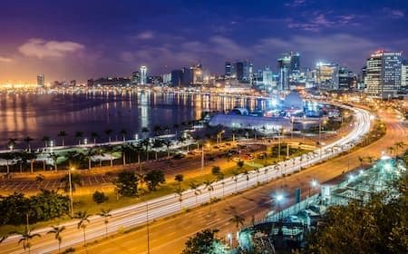 Nighttime aerial view of a coastal city skyline with illuminated buildings, waterfront, and highway traffic.