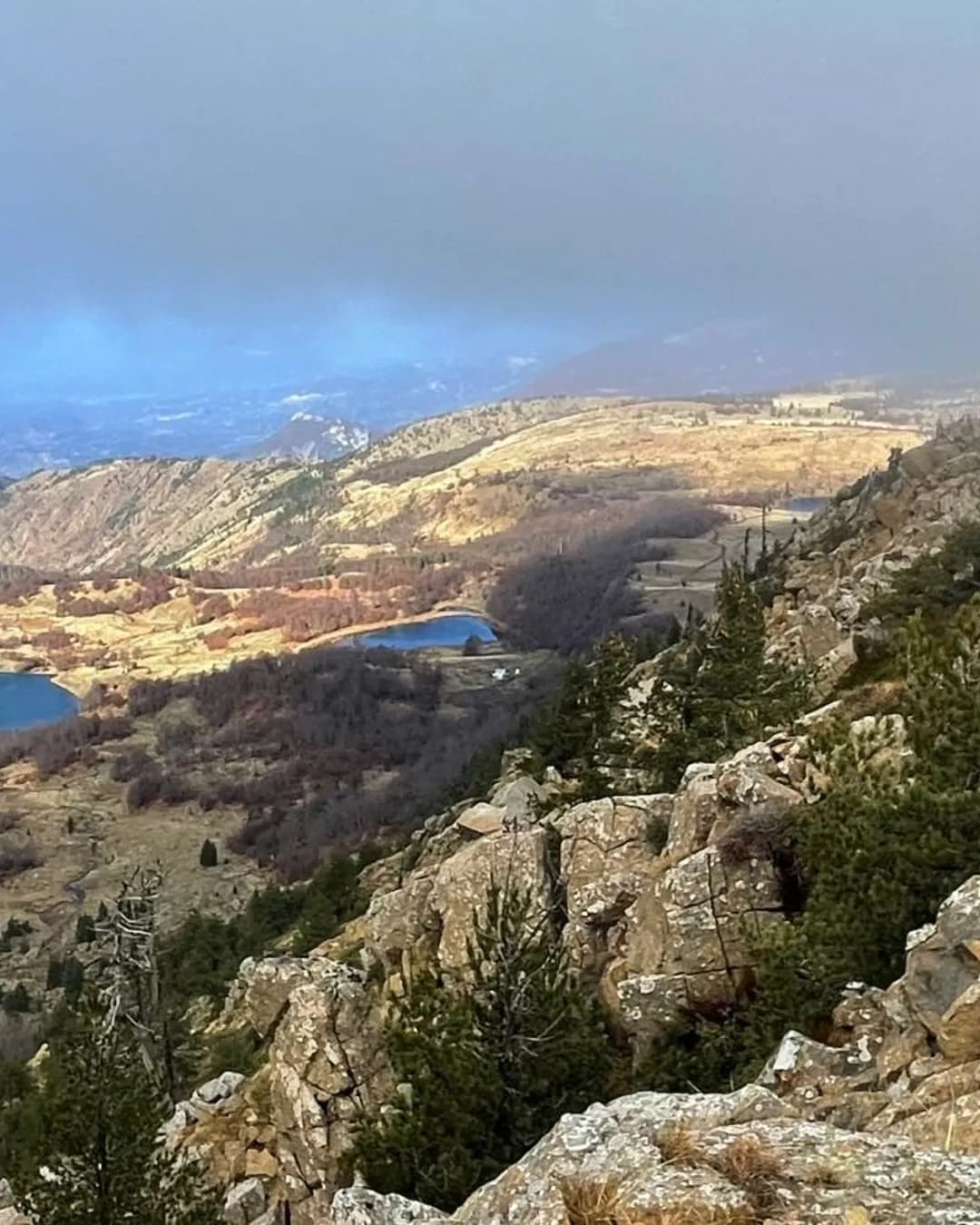 Scenic mountain landscape with rocky foreground, pine trees, rolling hills, and small blue lakes in a valley under a hazy sky.