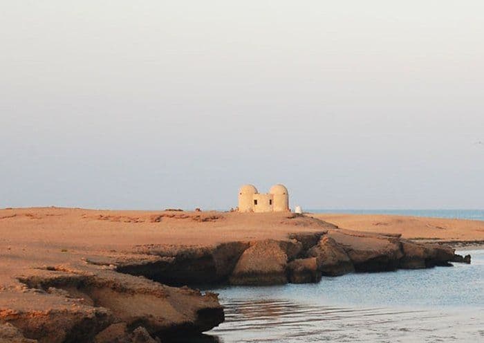Wide arid coastal landscape with sandy terrain, rocky shoreline, calm water, and two small domed structures on the horizon at Kamaran.