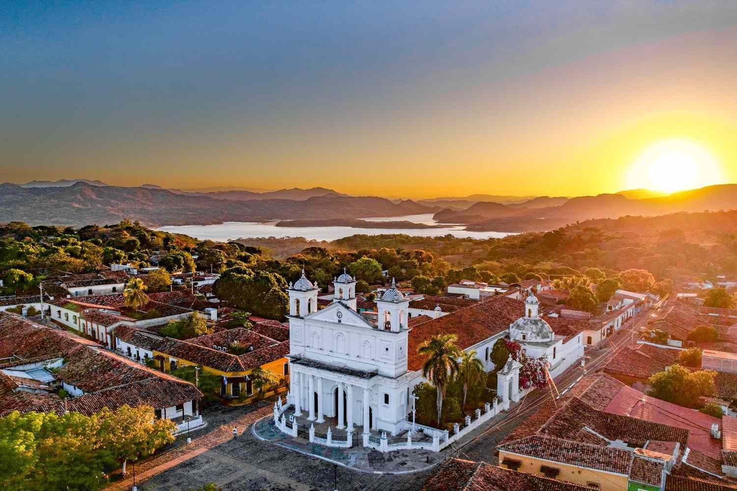 Aerial sunset view of a white colonial church with twin bell towers, surrounded by red-roofed buildings, plaza, and distant lake and mountains