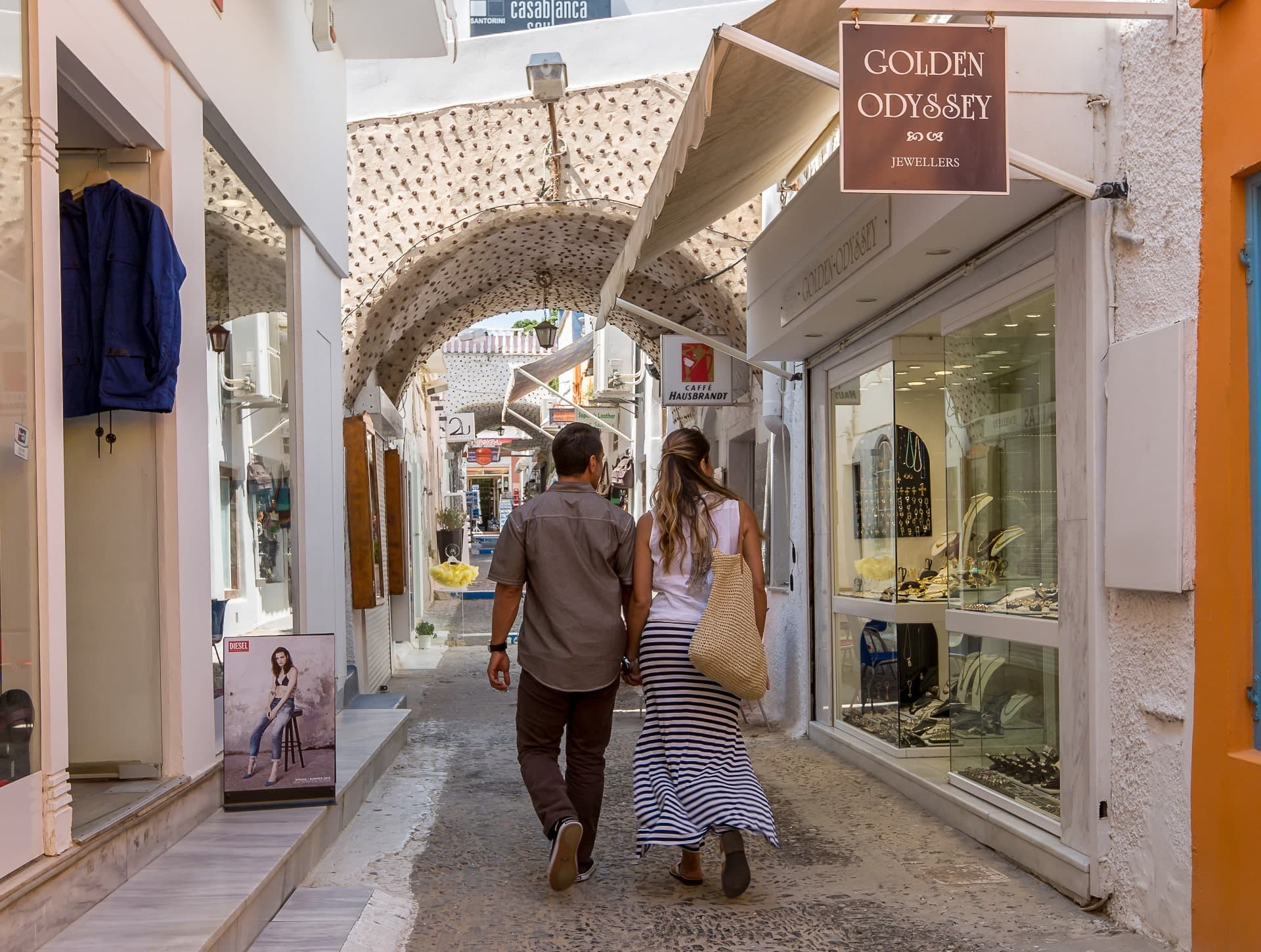 Couple walking away down a narrow, whitewashed street in Firá, passing shops like Golden Odyssey Jewellers under a textured arched ceiling.