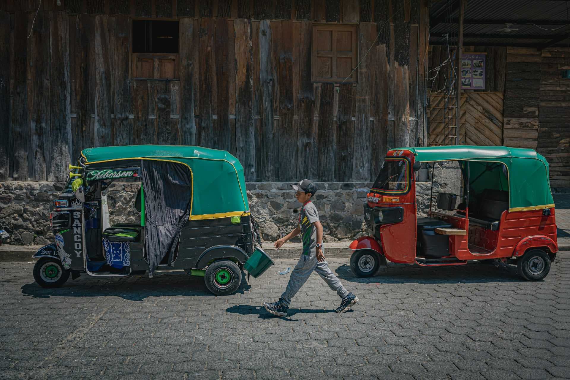 Two colorful tuk-tuks parked on a cobblestone street with a wooden building background; a boy walks between them carrying a bucket.