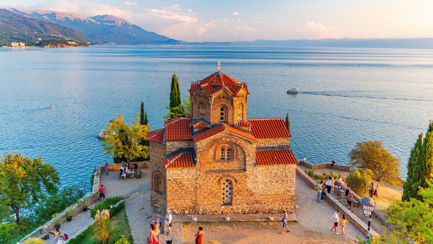 Aerial view of a stone church with red roofs on a lakeside terrace, overlooking Lake Ohrid and distant mountains.