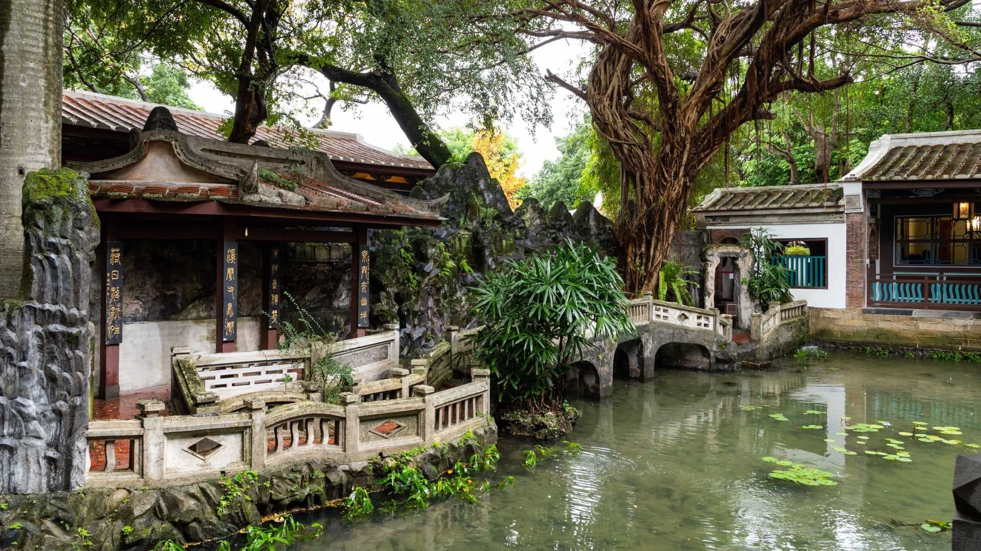 Traditional Chinese garden featuring a tranquil pond, stone bridges, and historic pavilions with intricate roof details, likely the Lin Family Mansion.