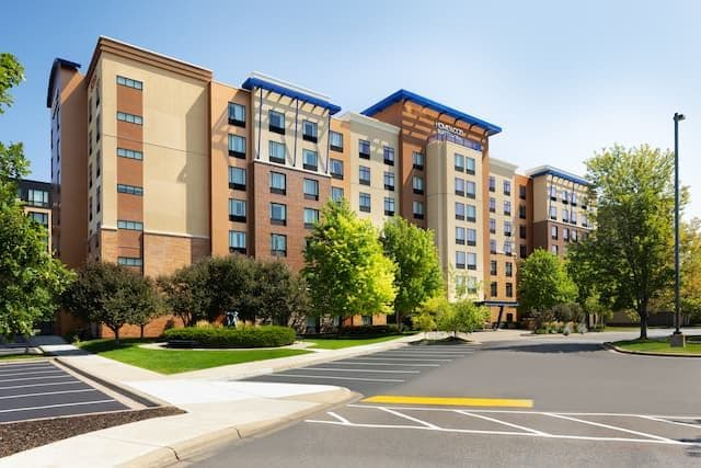Exterior of a modern mid-rise hotel building with brick and stucco facade, blue accents, parking lot, and trees.