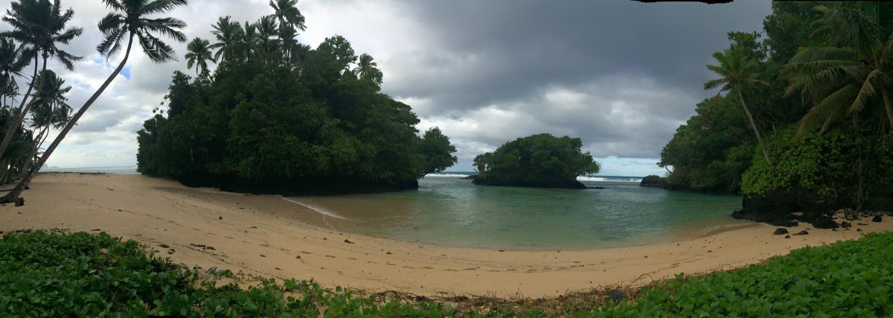 Coastal panorama of the Samamea shoreline with sandy beach, palm trees, lush vegetation, and calm turquoise water under a cloudy sky.