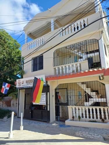 A street-level view of a multi-story building with balconies and flags, situated on a paved road in a small town.
