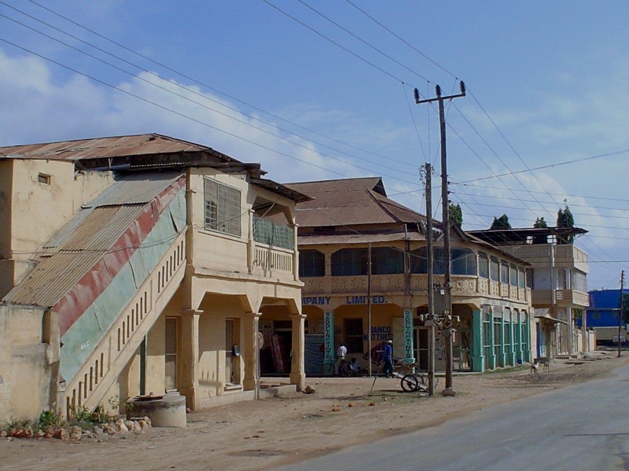 Street view of two-story buildings with shops and a staircase in Lindi, Tanzania, under a blue sky.
