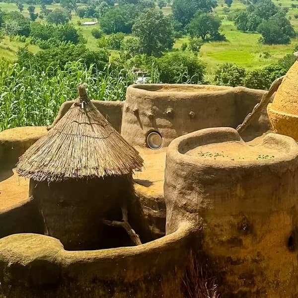 Traditional earthen architecture with rounded mud structures and a thatched conical roof, set against a green rural hillside with trees and crops