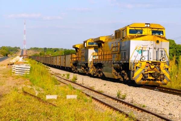 A yellow diesel locomotive pulls a long freight train along railway tracks through a grassy landscape under a blue sky.