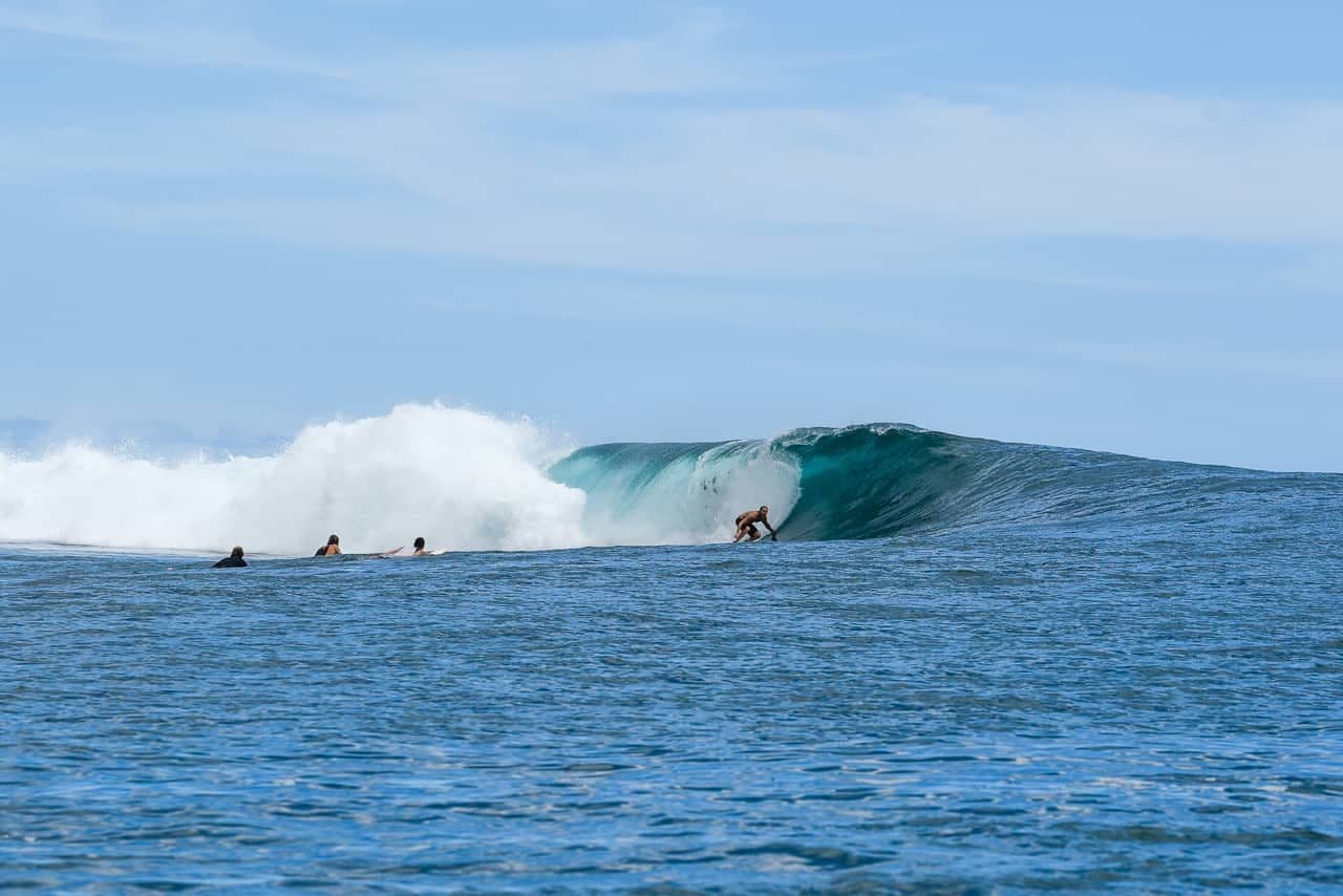 Surfers ride and wait on a large breaking wave in open blue ocean under a clear sky near Samamea, Samoa.