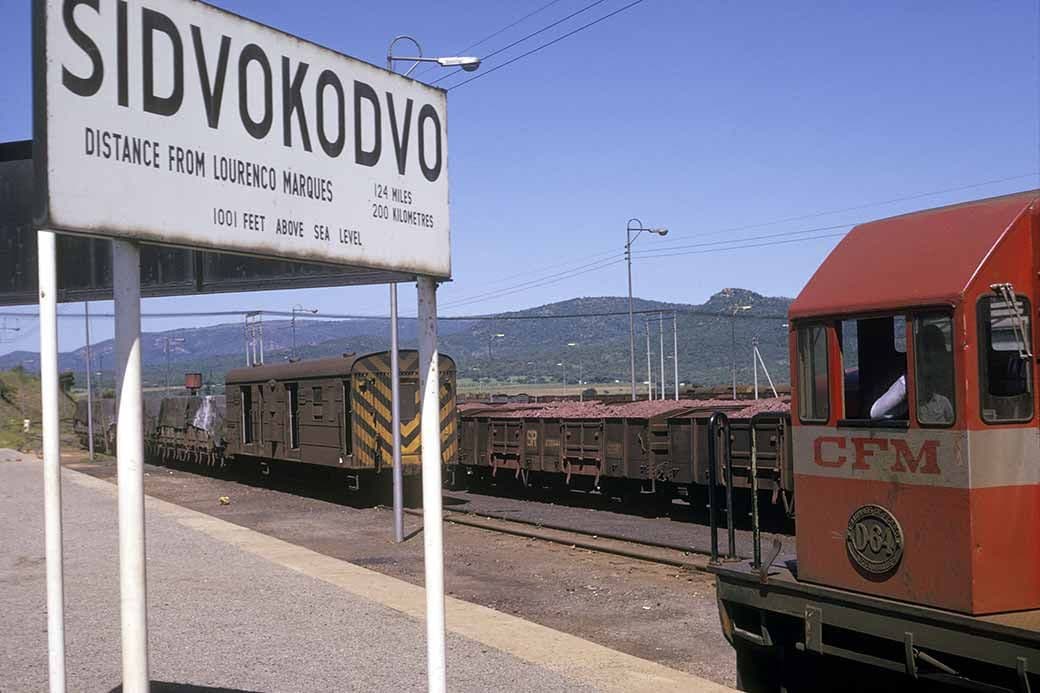 Railway station sign reading Sidvokodvo, freight wagons on tracks, and a red CFM railcar under a clear sky.