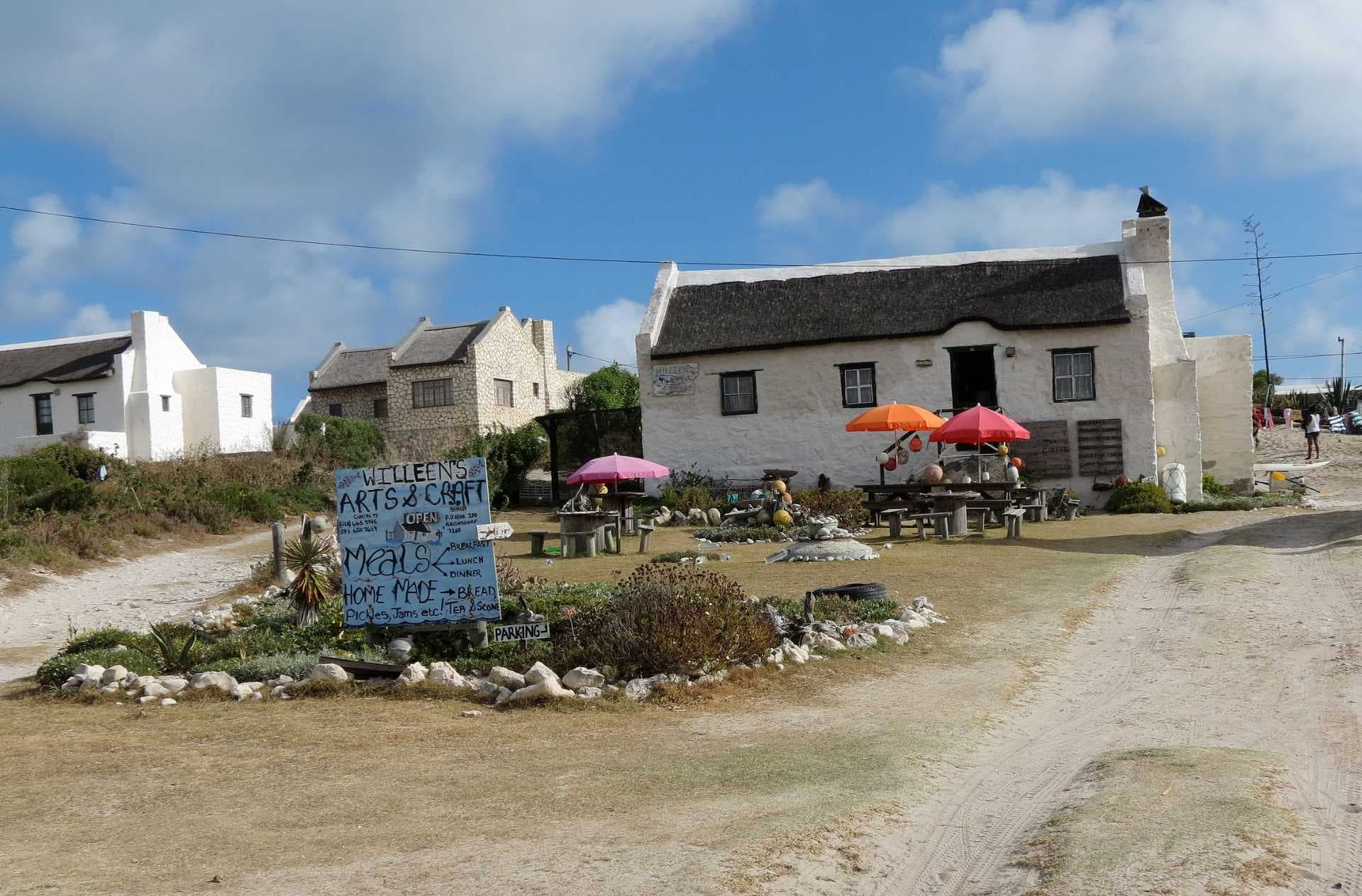 Village street with white thatched-roof houses, outdoor dining umbrellas, and a hand-painted meals sign under a blue sky.