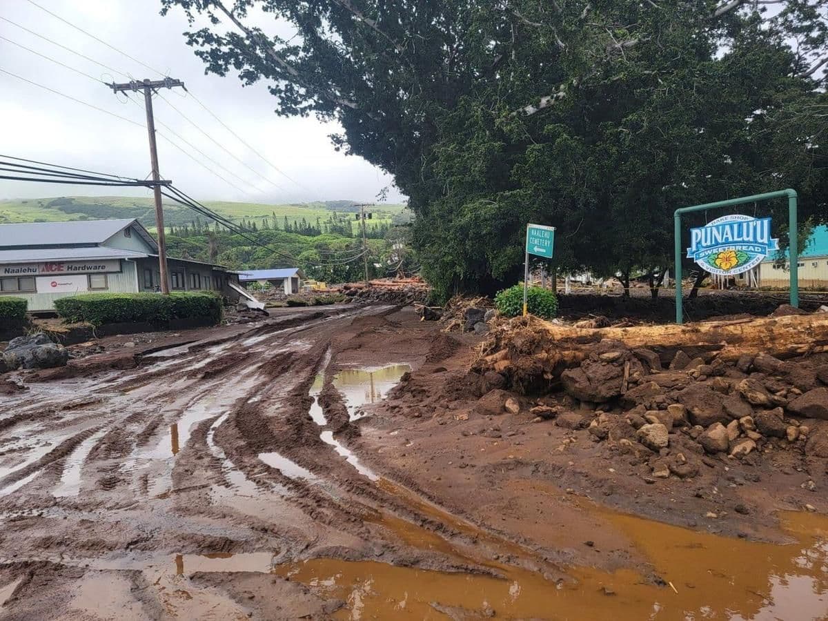 Muddy, flooded street in a small town with local shops, signage, and green hills in the background near Mengomeyén, Equatorial Guinea