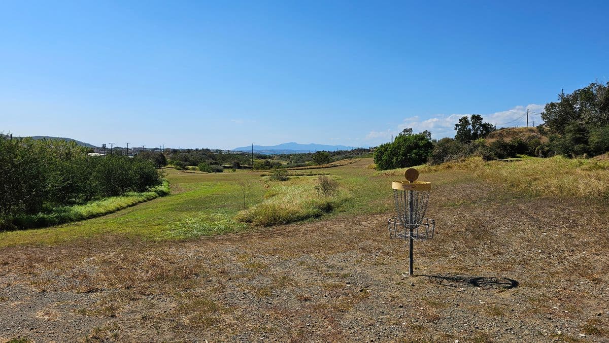 Rural open field with a disc golf basket, dry grass, distant hills, and clear blue sky.