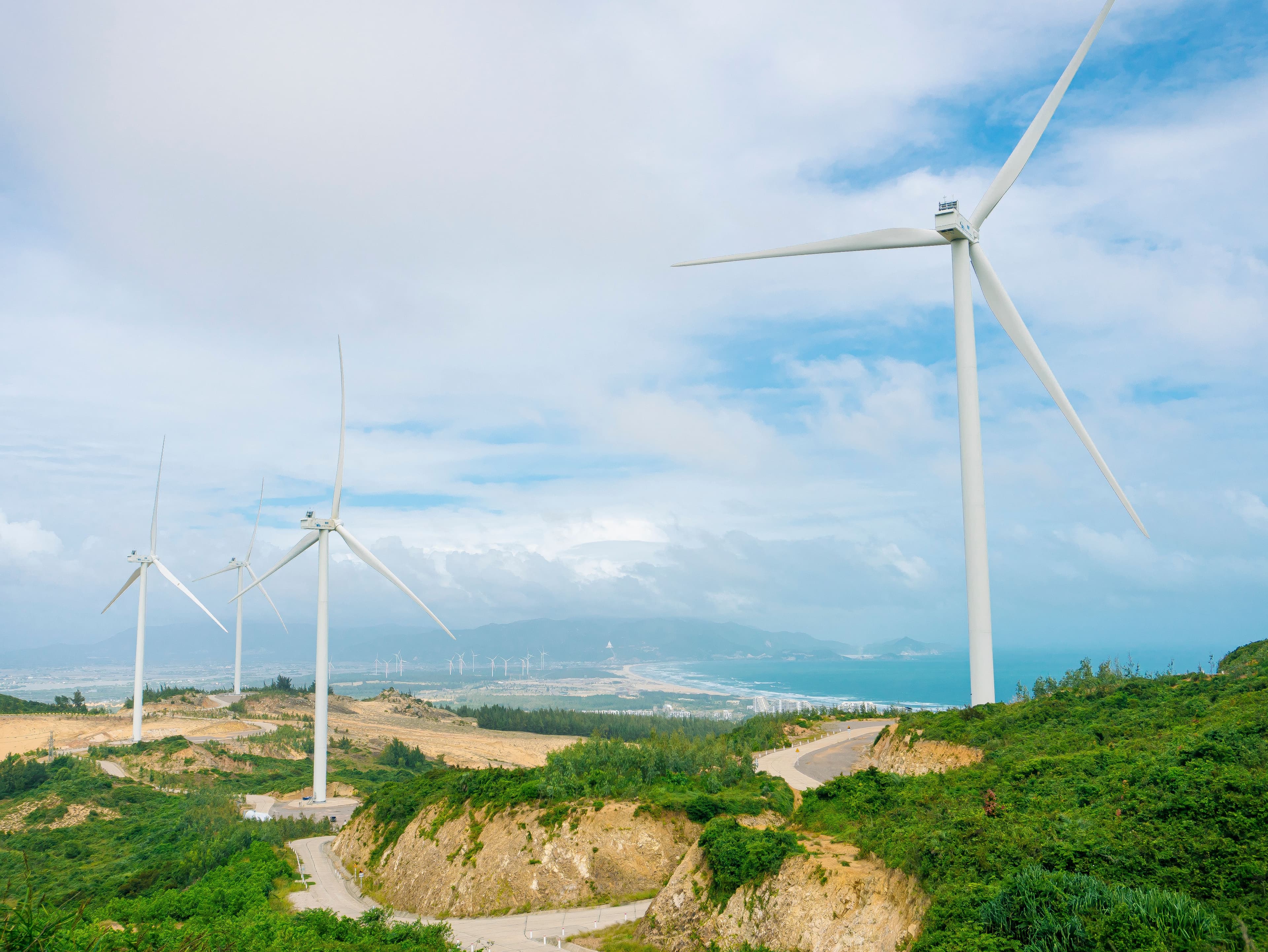 Winding paved road traverses a green hillside dotted with large white wind turbines, overlooking a distant coastline and ocean under a cloudy sky.