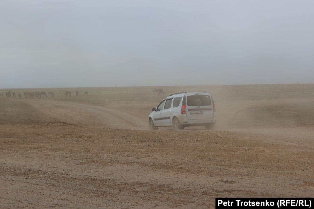 A white van drives across a dusty, open steppe landscape near the Kazakhstan–Russia border with distant livestock under an overcast sky.