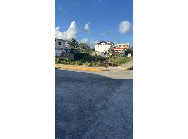 Street-level view of a paved road corner with yellow curbs, overlooking a grassy lot and residential buildings under a blue sky with clouds.