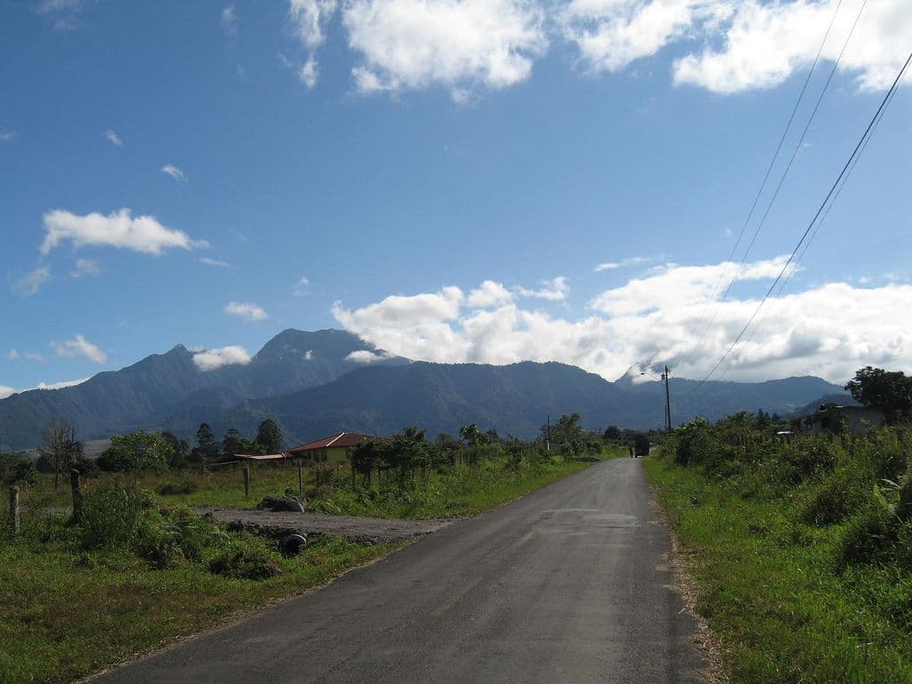 Paved rural road stretches toward green mountains under a partly cloudy blue sky in the highlands near Mirono, Panama.