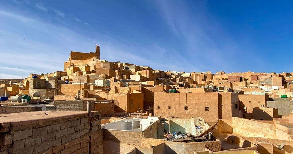 Wide exterior view of a hillside kasbah town with earthen buildings under a clear blue sky in the Ghardaia province near Smala.