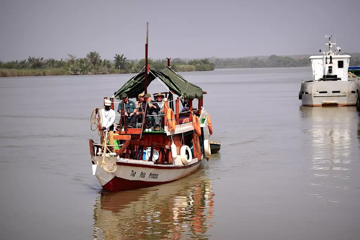 A small wooden passenger boat with a green canopy travels on a wide river, with a larger vessel and distant shoreline visible.