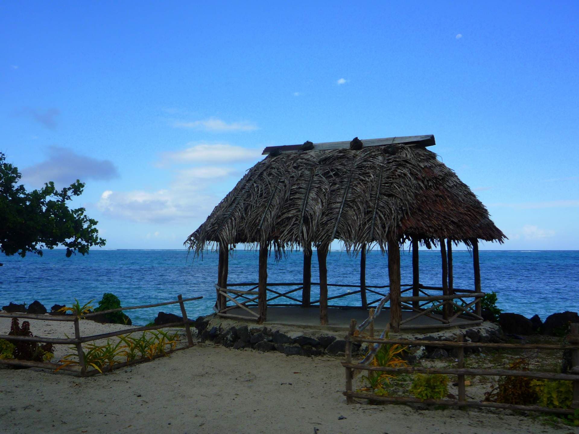 A thatched-roof open pavilion stands on a sandy beach with the ocean in the background in Samoa.
