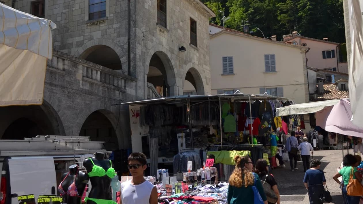 Outdoor market stalls selling clothing and swimwear in a European town square, with pedestrians and historic stone architecture