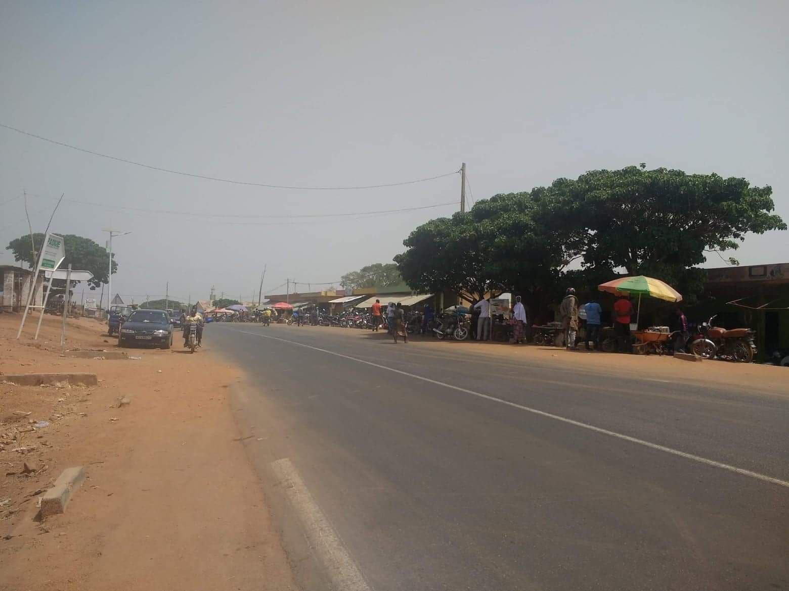 Wide street scene in Bembèrèkè with roadside market stalls, parked motorcycles, a large tree, and a hazy sky.