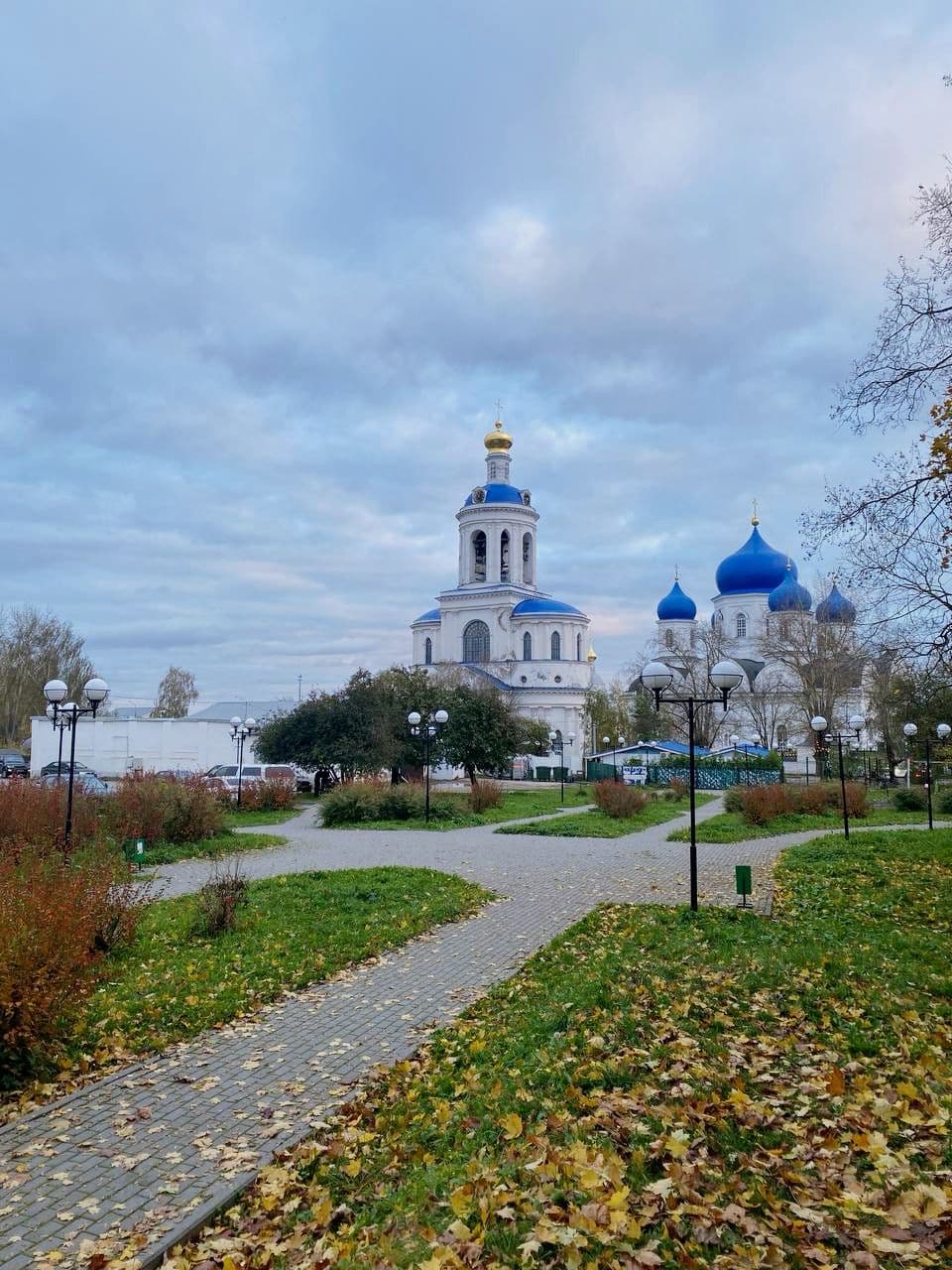 Paved park path leading to white Orthodox churches with blue domes under an overcast sky in the Tula Oblast region near Tonshalovo.