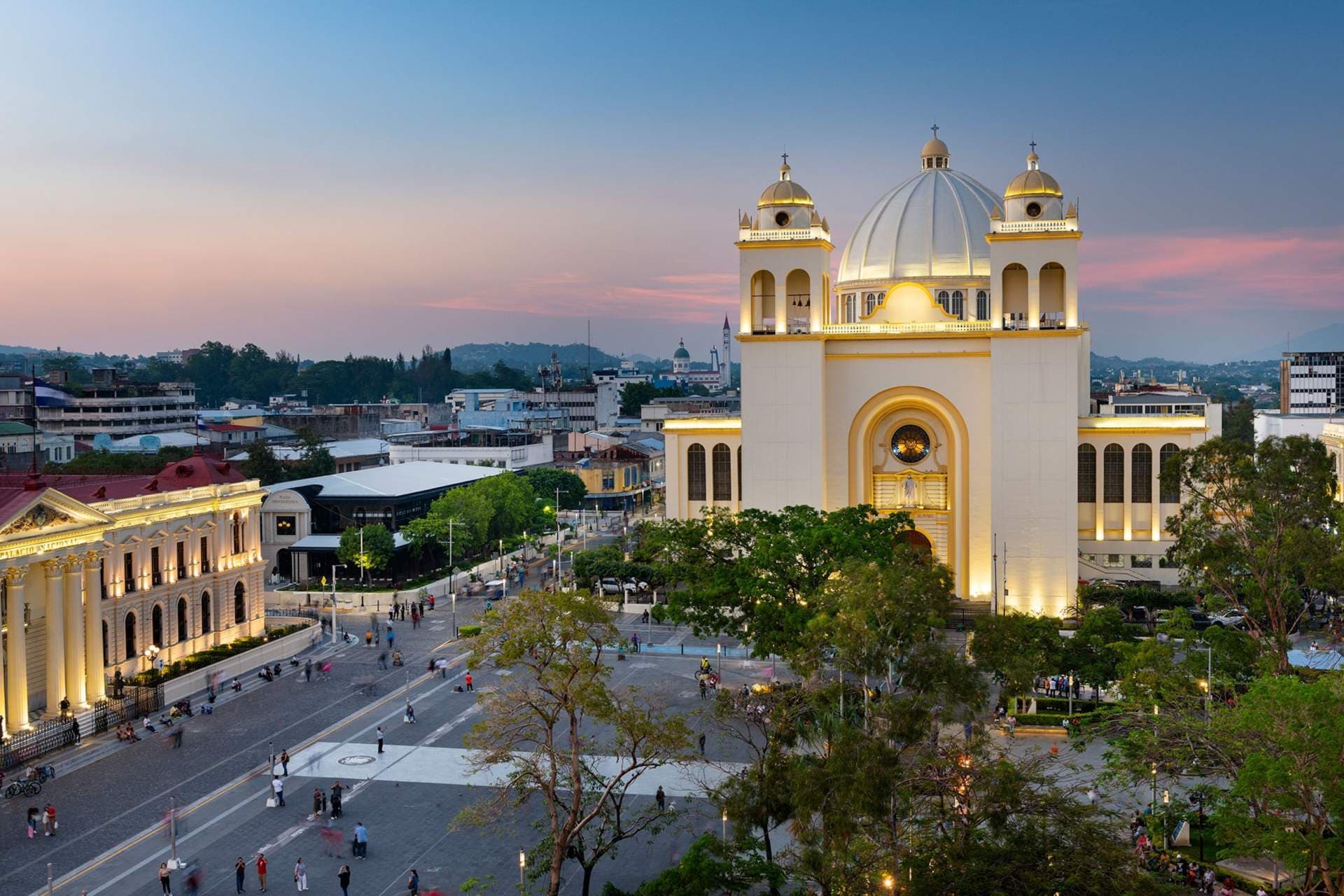 Aerial dusk view of a large white cathedral with twin towers and dome, adjacent to a plaza in El Salvador.