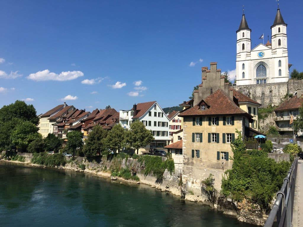 Traditional Swiss village buildings and a twin-towered church line the riverbank under a clear blue sky.
