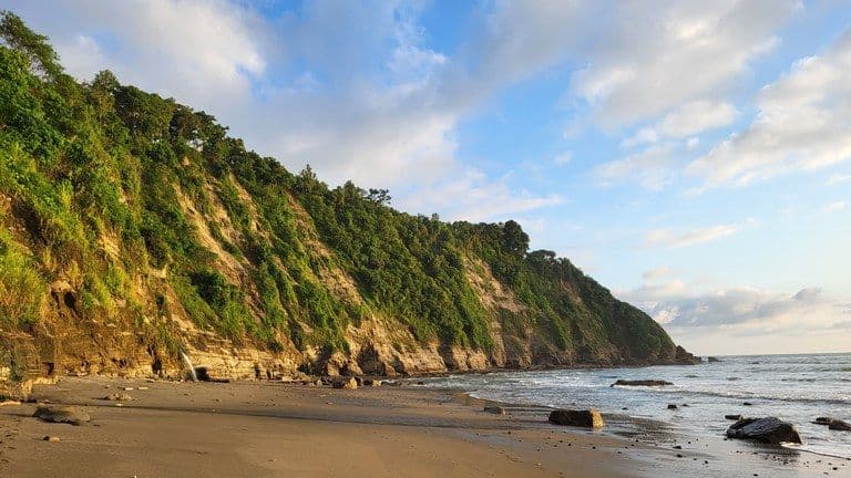 Coastal landscape near Pedernales, Ecuador, showing a sandy beach, rocky shoreline, and green cliffs meeting the Pacific Ocean under a partly cloudy sky.