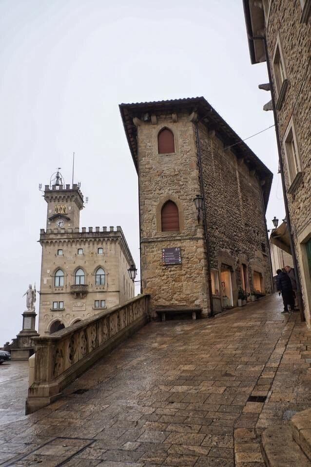 Wet stone-paved street in an old town with historic stone buildings and a clock tower under an overcast sky
