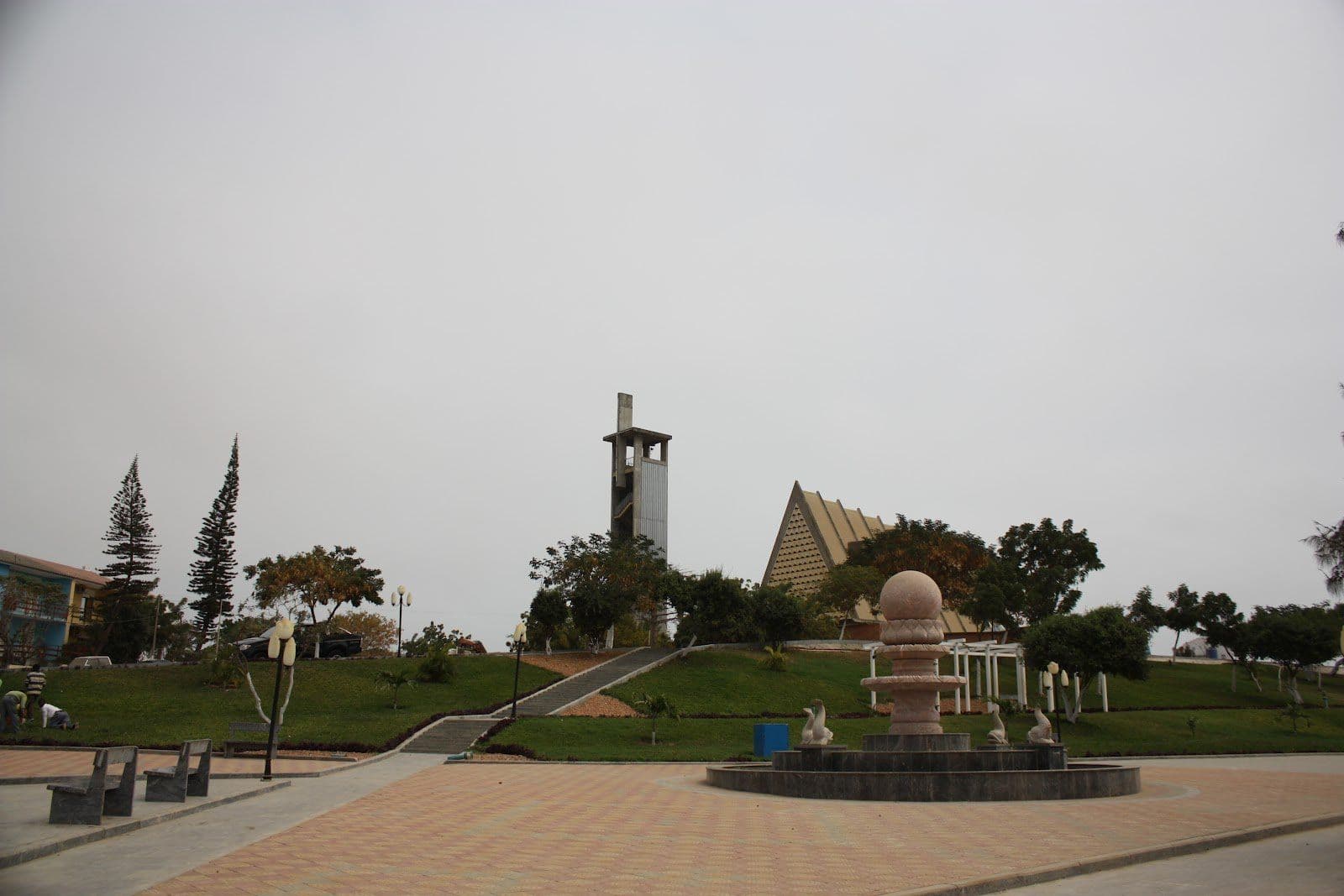 Wide view of a paved public plaza with a stone fountain, manicured lawns, benches, and a modern church with a tall bell tower under an overcast sky.