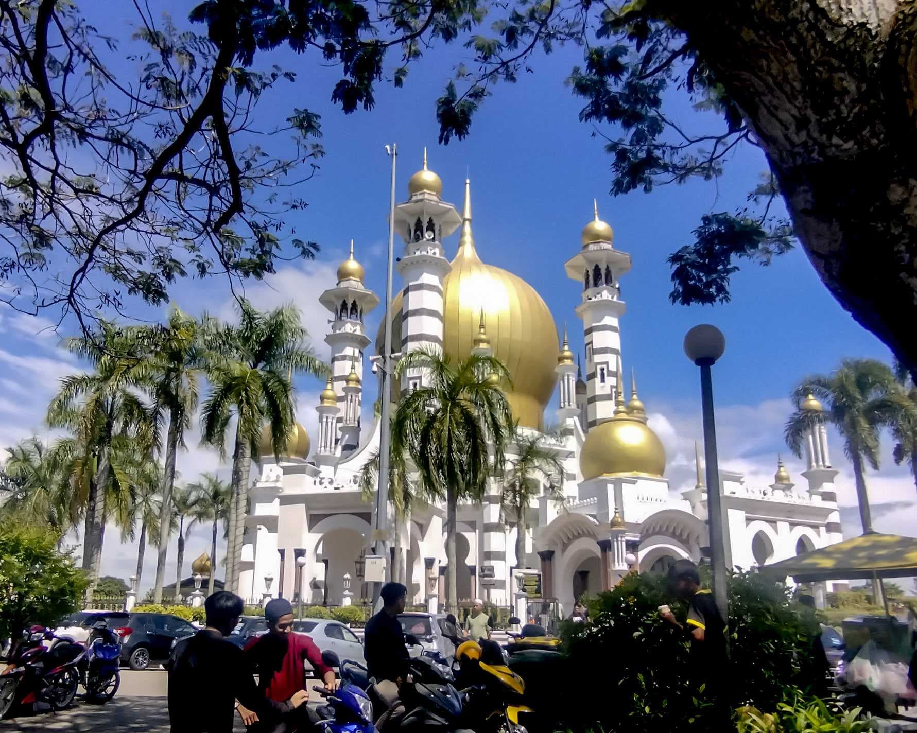 Exterior of Ubudiah Mosque with golden domes, white minarets, palm trees, and parked motorcycles under a blue sky