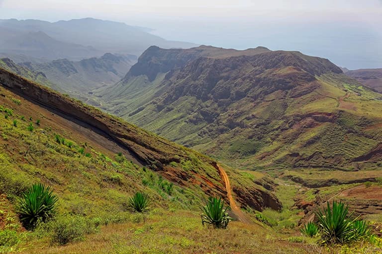 A scenic view of the rugged, arid interior of Santiago, featuring steep green and brown hills, a winding dirt road, and distant mountains.