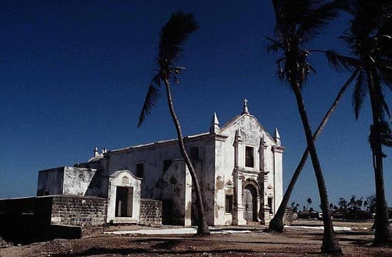 Exterior view of a weathered white historic building with a cross and palm trees against a deep blue sky.