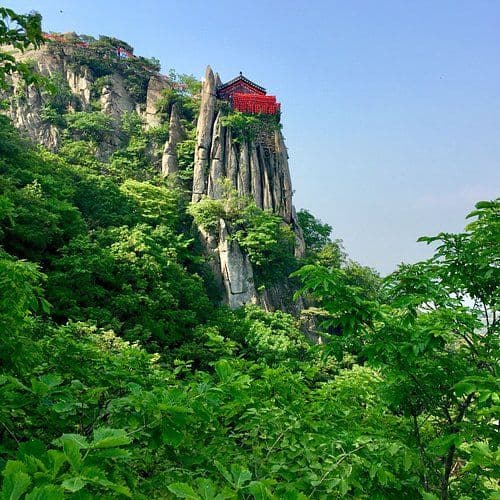 Rocky mountain slope with dense green foliage and a red pavilion perched on a cliff under a clear blue sky.