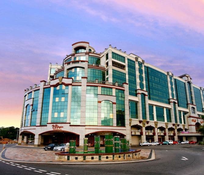 Large modern glass-fronted commercial building with curved facade, parked cars, and a small fountain plaza at dusk in Bandar Seri Begawan.
