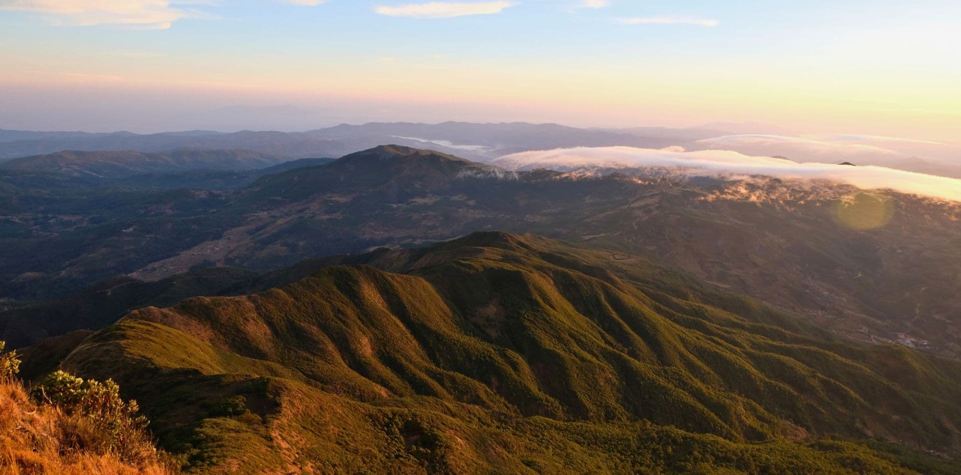 Wide highland landscape with scrub-covered ridges, open terrain, distant mountains, and low clouds at golden hour.