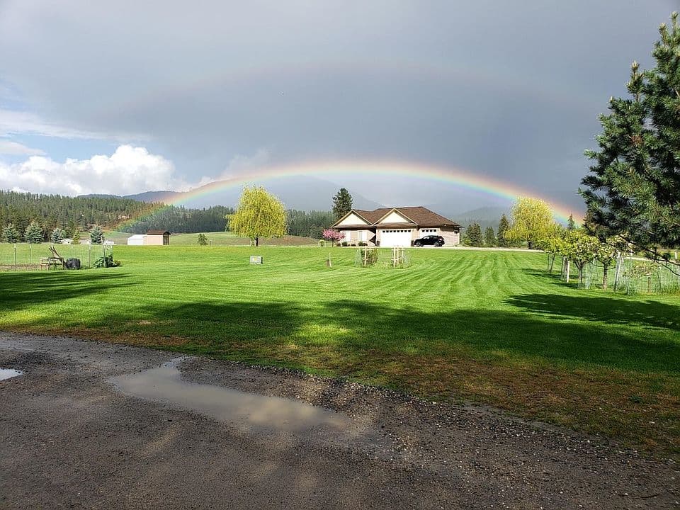 A scenic landscape of Chewelah, Washington, featuring a house with a rainbow arching over the roof against a backdrop of mountains and green fields.