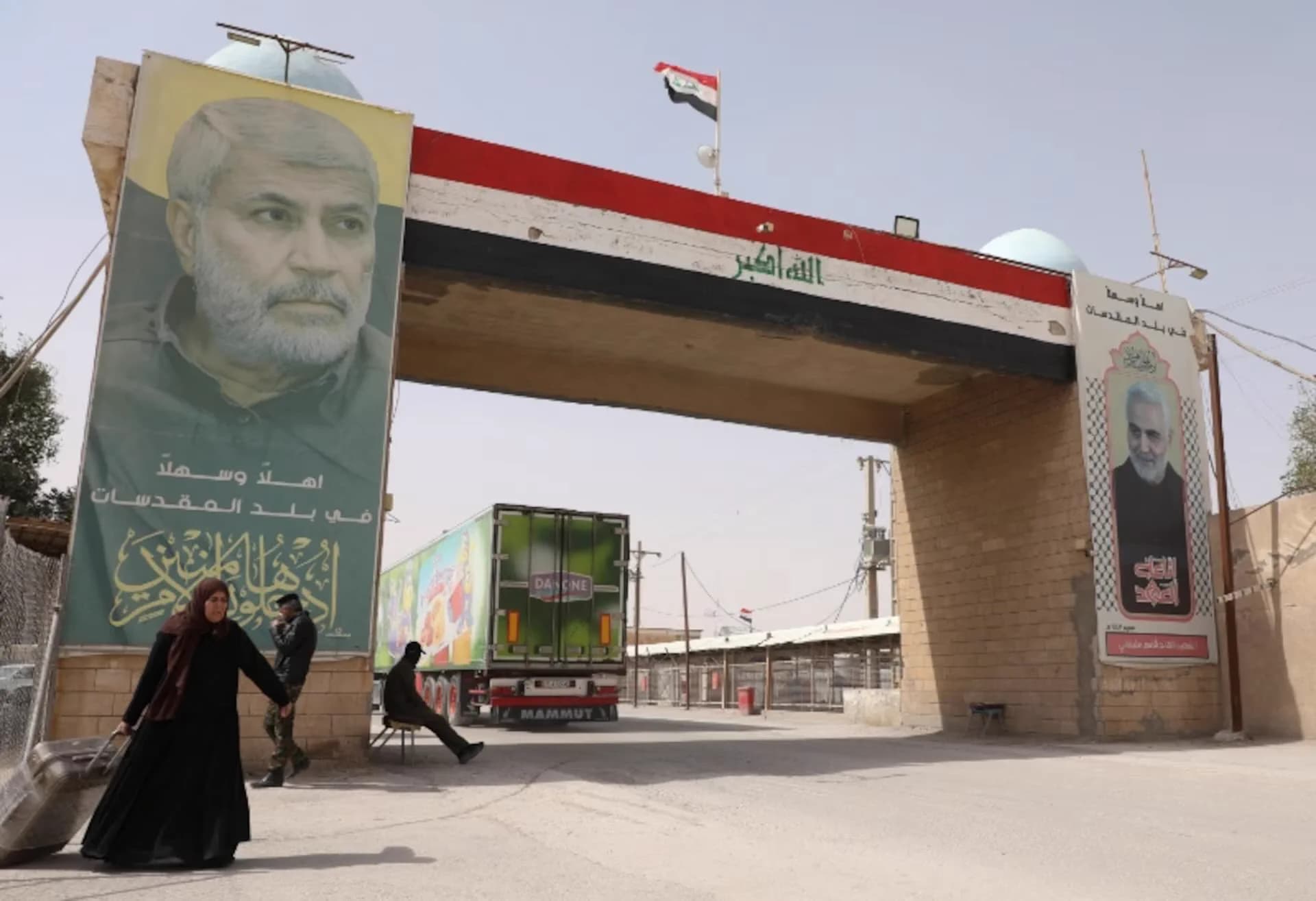 Large concrete archway with an Iraqi flag and political banners in Al ‘Amārah, with a truck and pedestrian in the background.